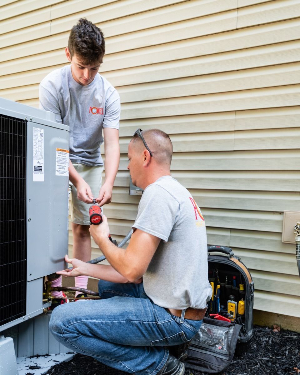 Two men are working on an air conditioner outside of a house.