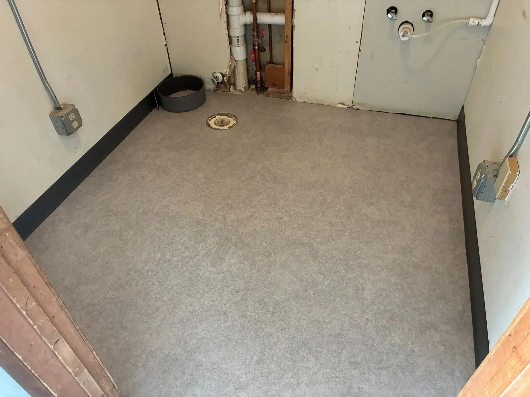 Small empty utility room with gray carpet, white walls, exposed pipes, and a drain on the floor.