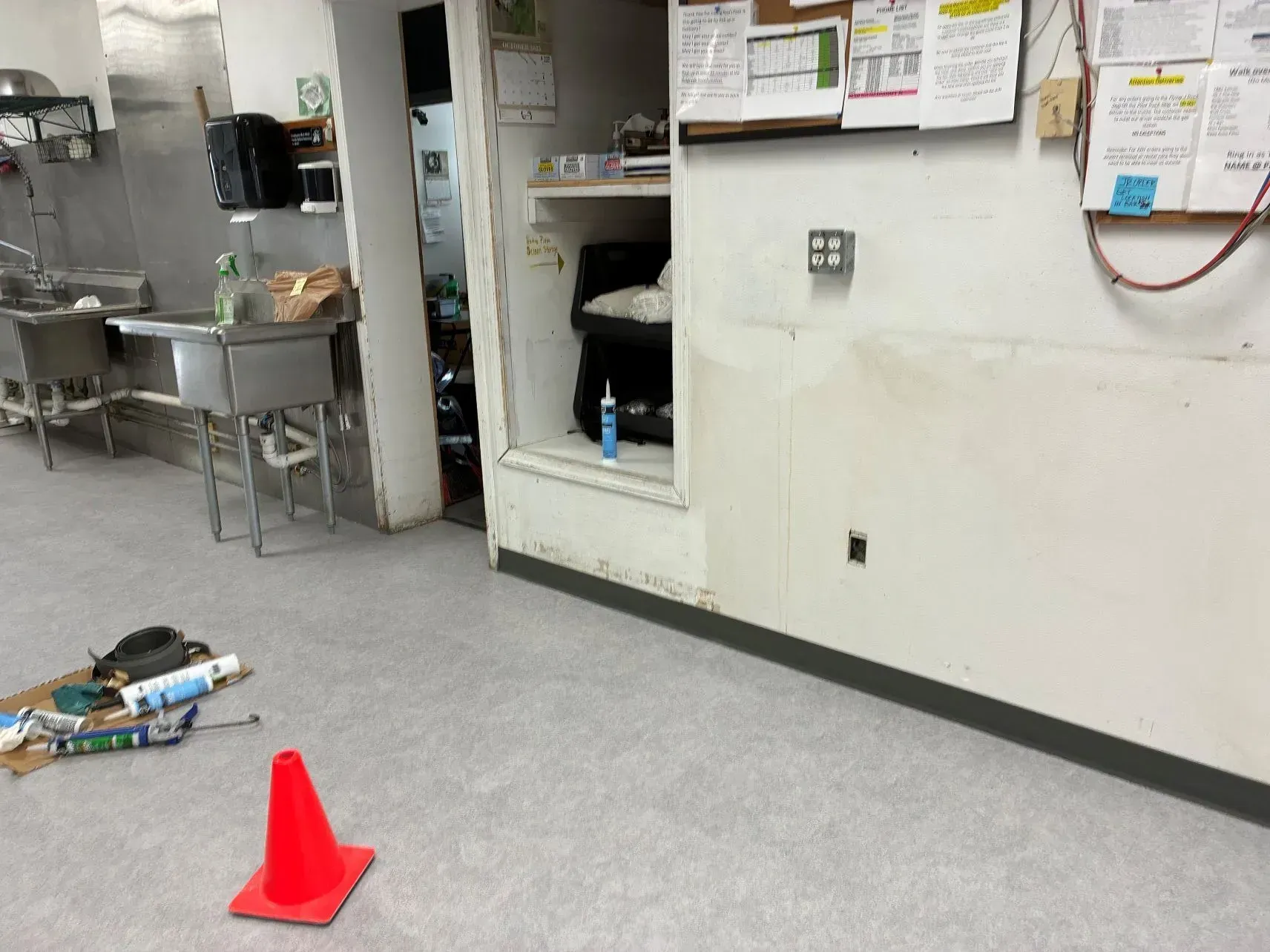 Industrial kitchen floor with orange cone and cleaning supplies near a wall opening.