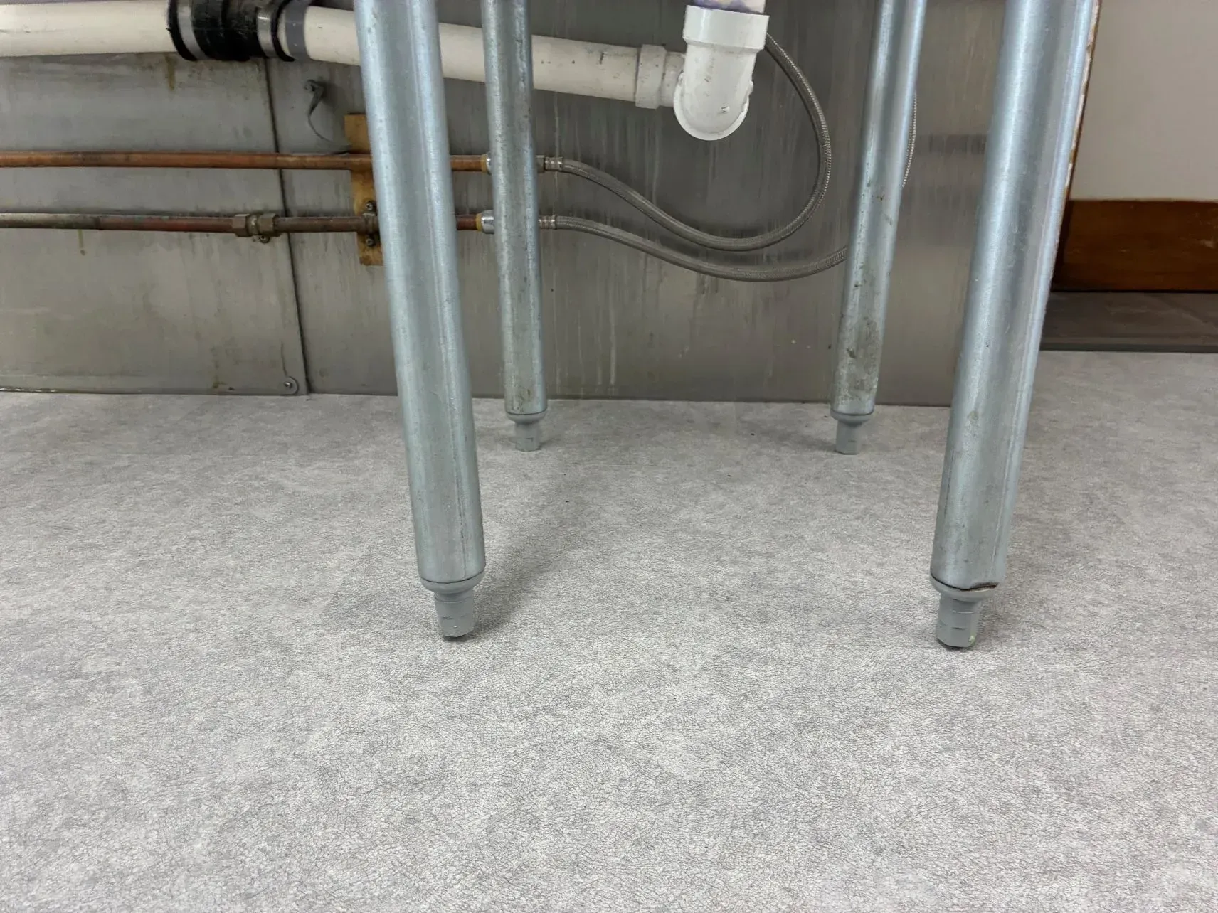 Metal table legs on a gray carpeted floor near exposed pipes and plumbing under a sink or counter.