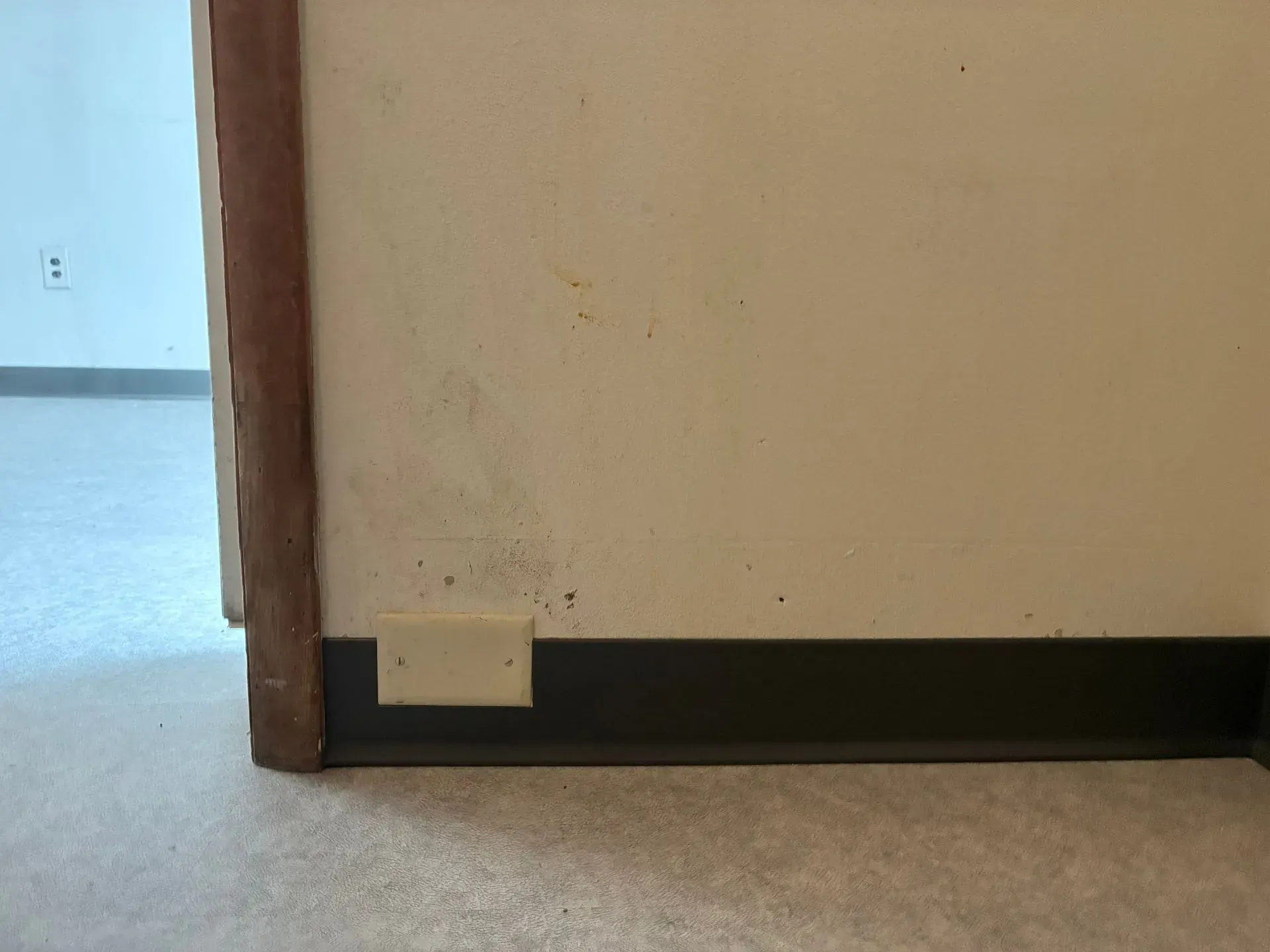 Corner of a beige hallway wall with a black baseboard and a small wall plate near the floor.