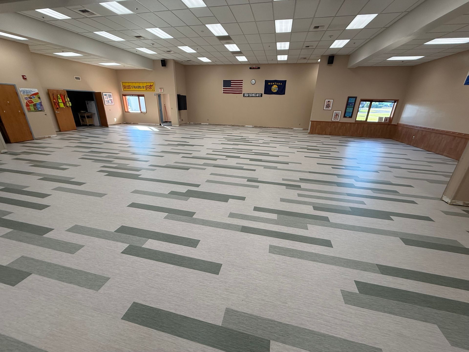 Empty school cafeteria with patterned gray carpet, beige walls, and bright ceiling lights.