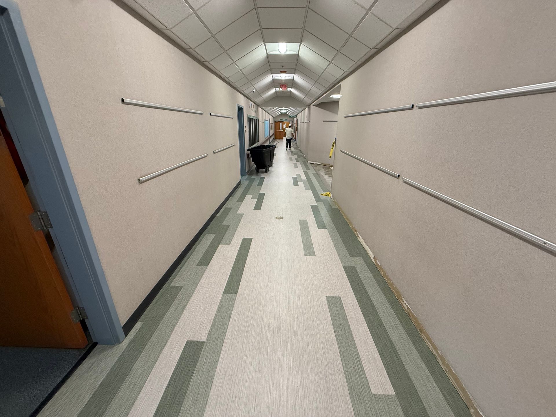 Long hallway with green and white tiled floor, beige walls, and a person at the far end.