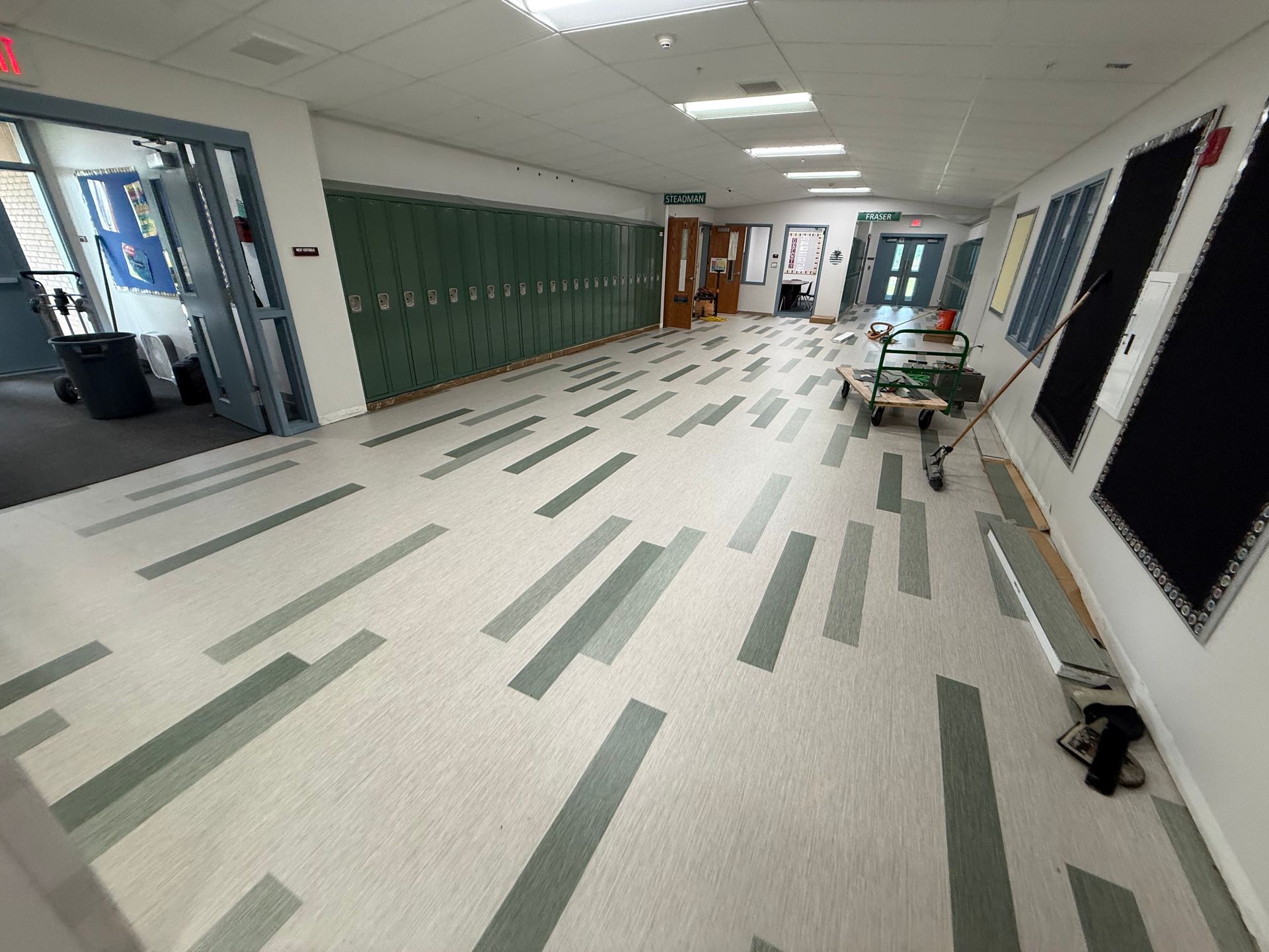 Empty school hallway with green lockers, white walls, and patterned tile floor; a cart sits near the windows.