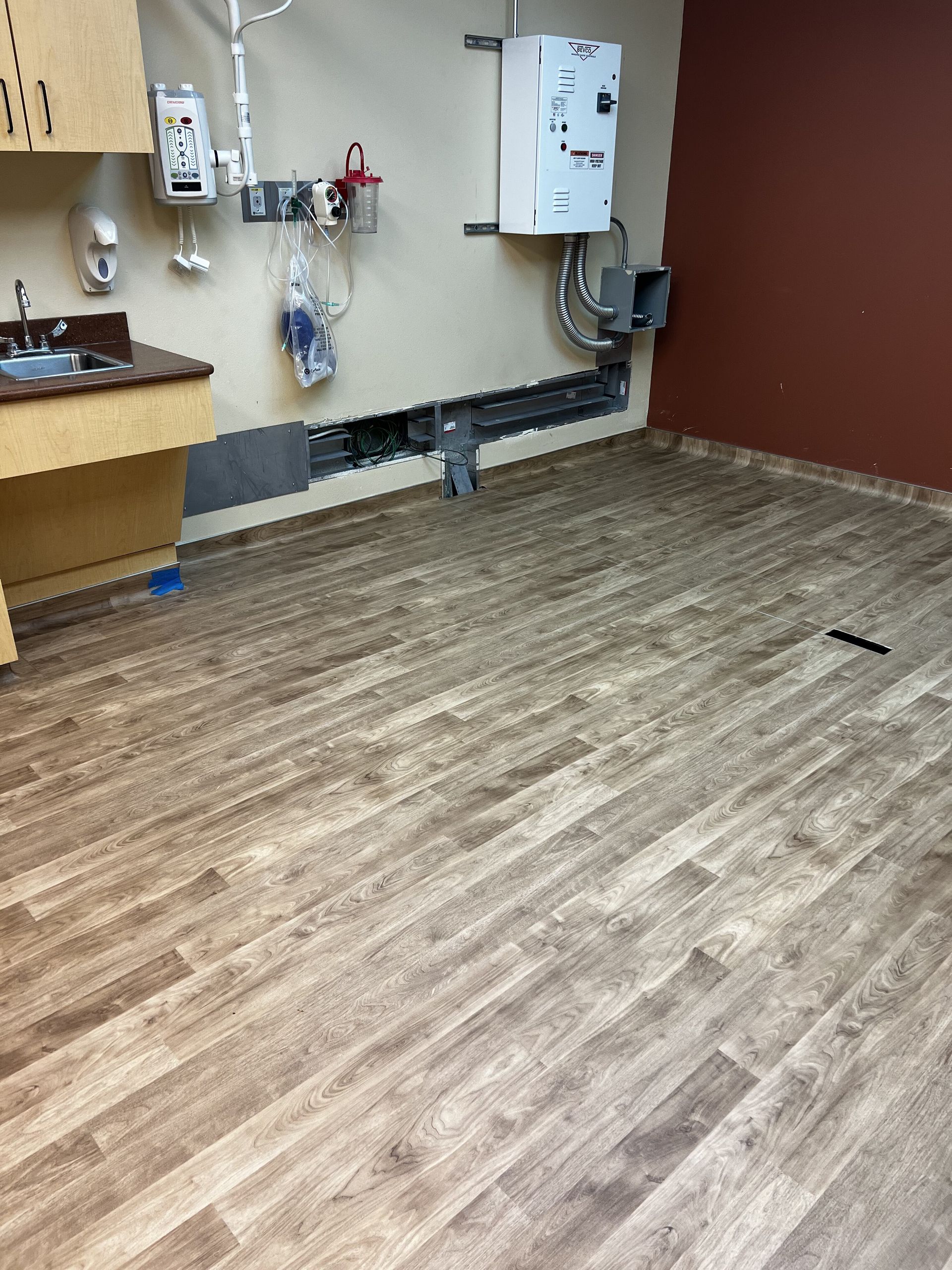 Empty clinic room with beige walls, medical equipment on the wall, and wood-look vinyl flooring.