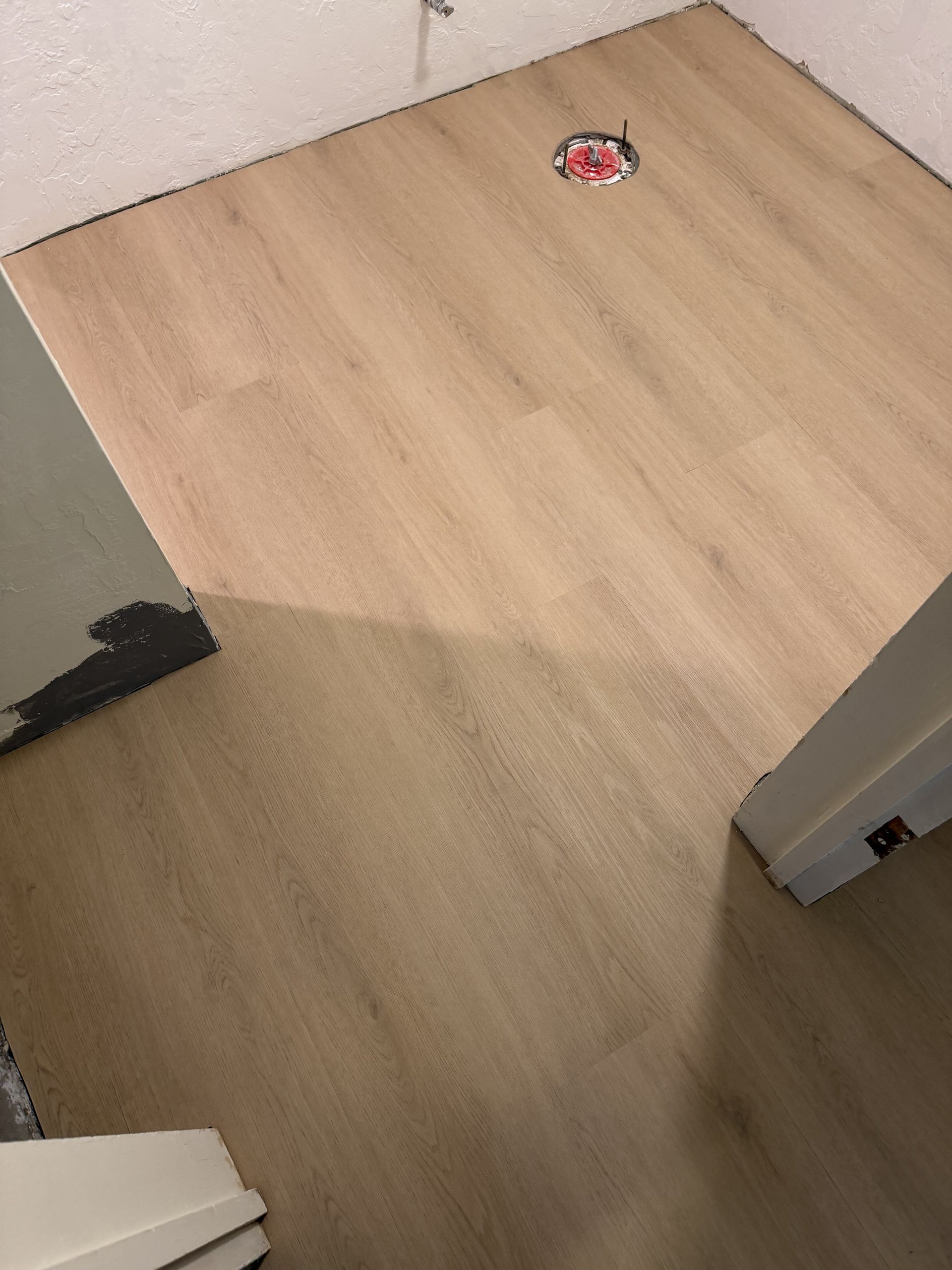 Small empty room corner with light wood flooring and a floor drain or vent near the wall.