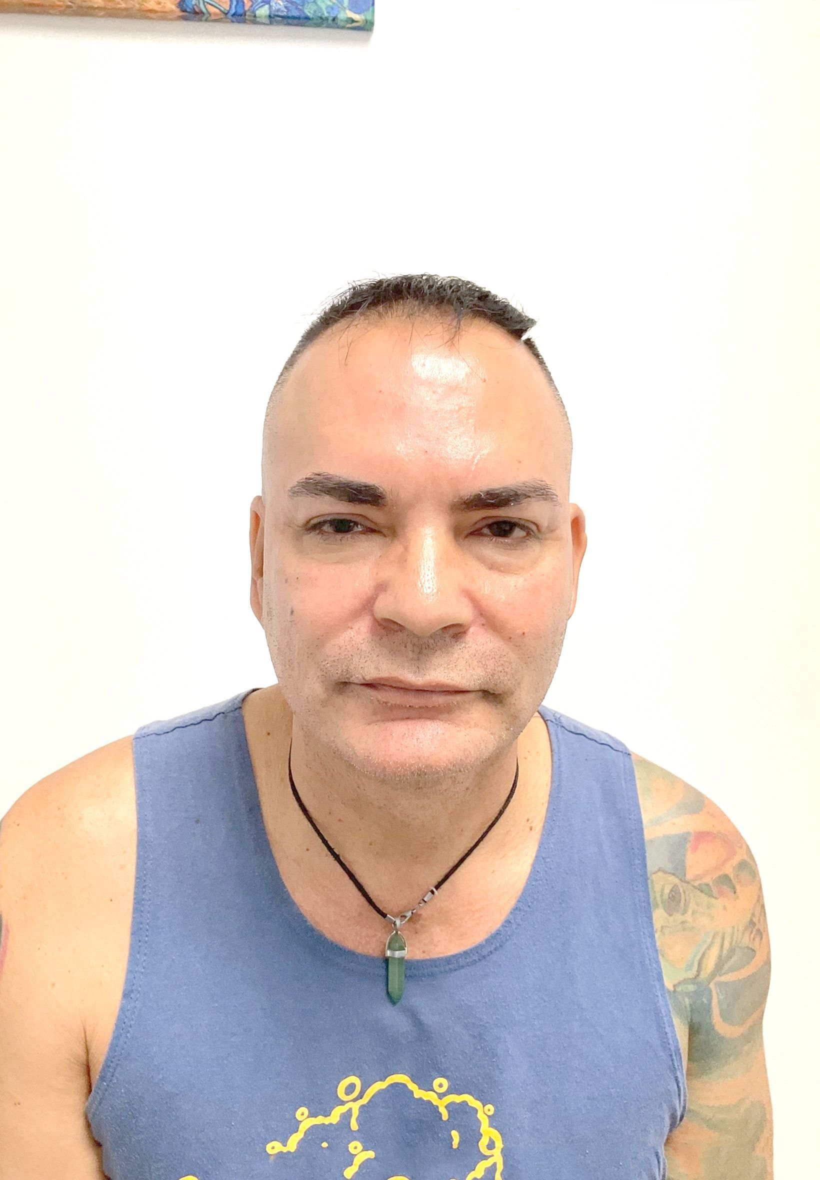 A man wearing a blue tank top and a necklace is standing in front of a white wall