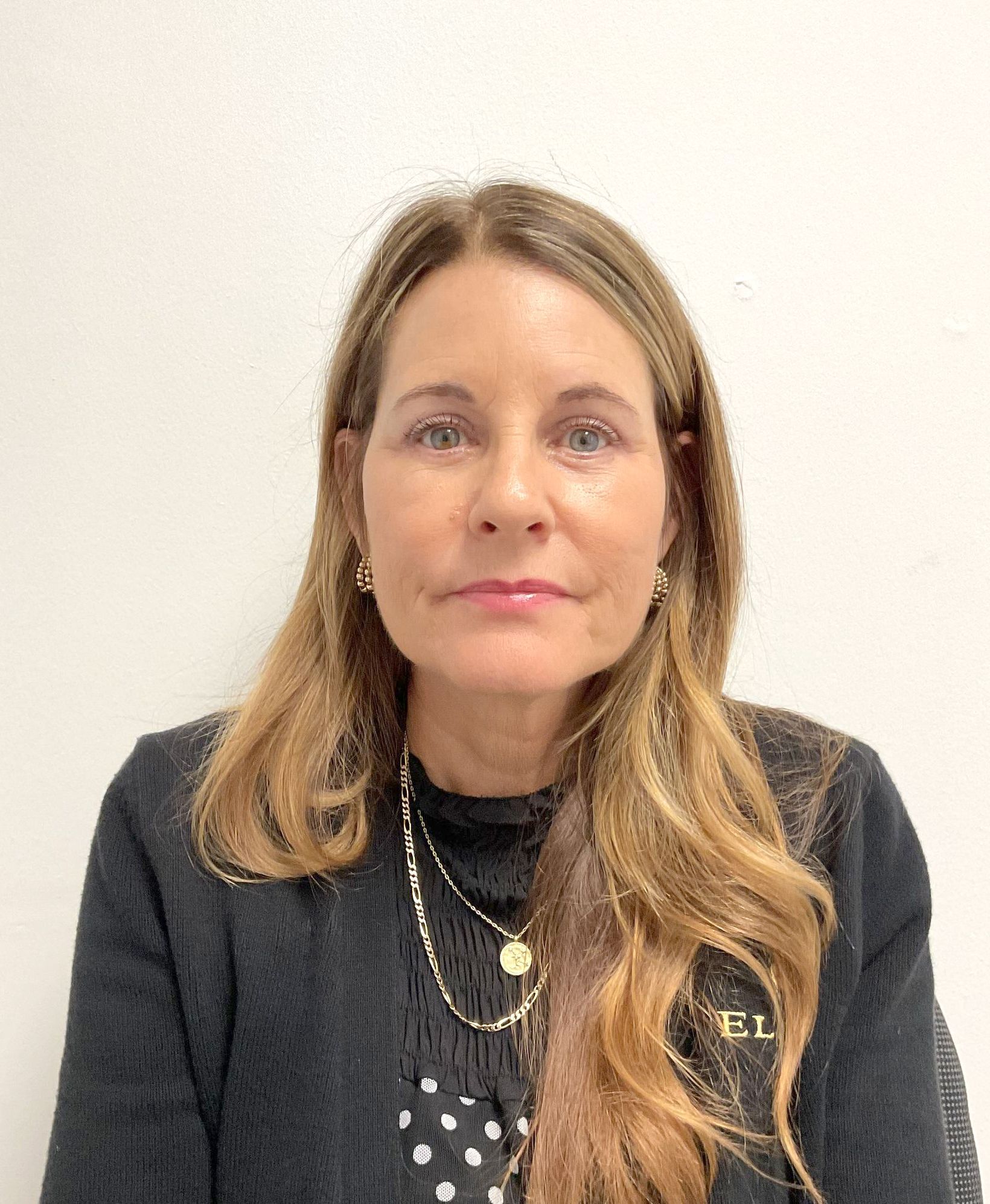 A woman wearing a black sweater and a polka dot shirt is sitting in front of a white wall
