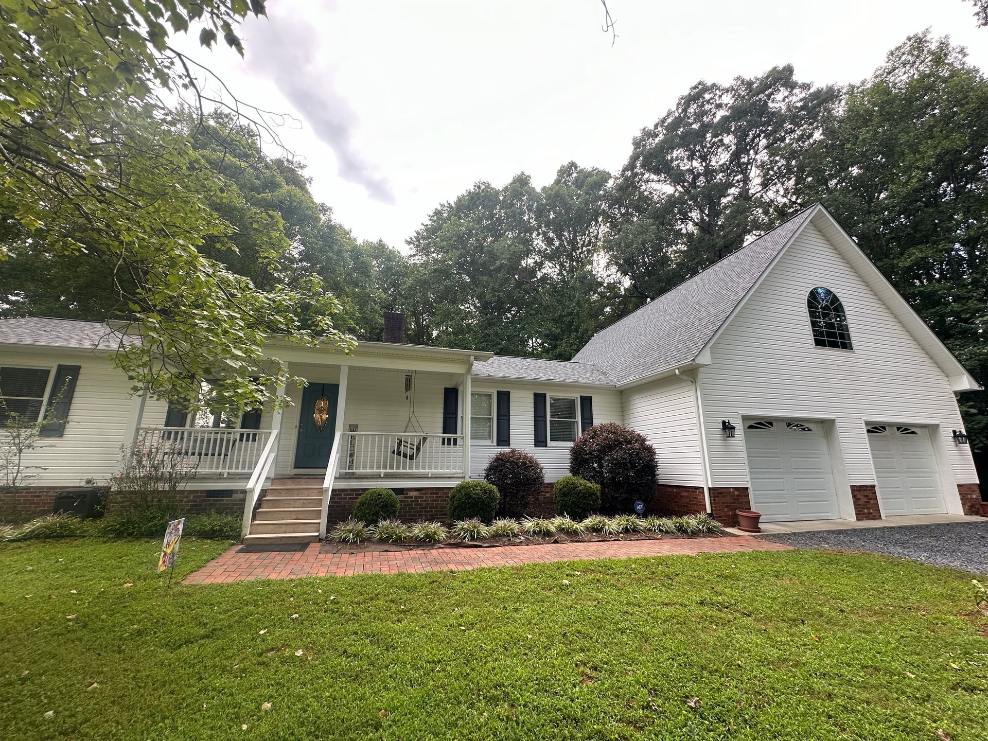 A white house with a porch and two garages is surrounded by trees.