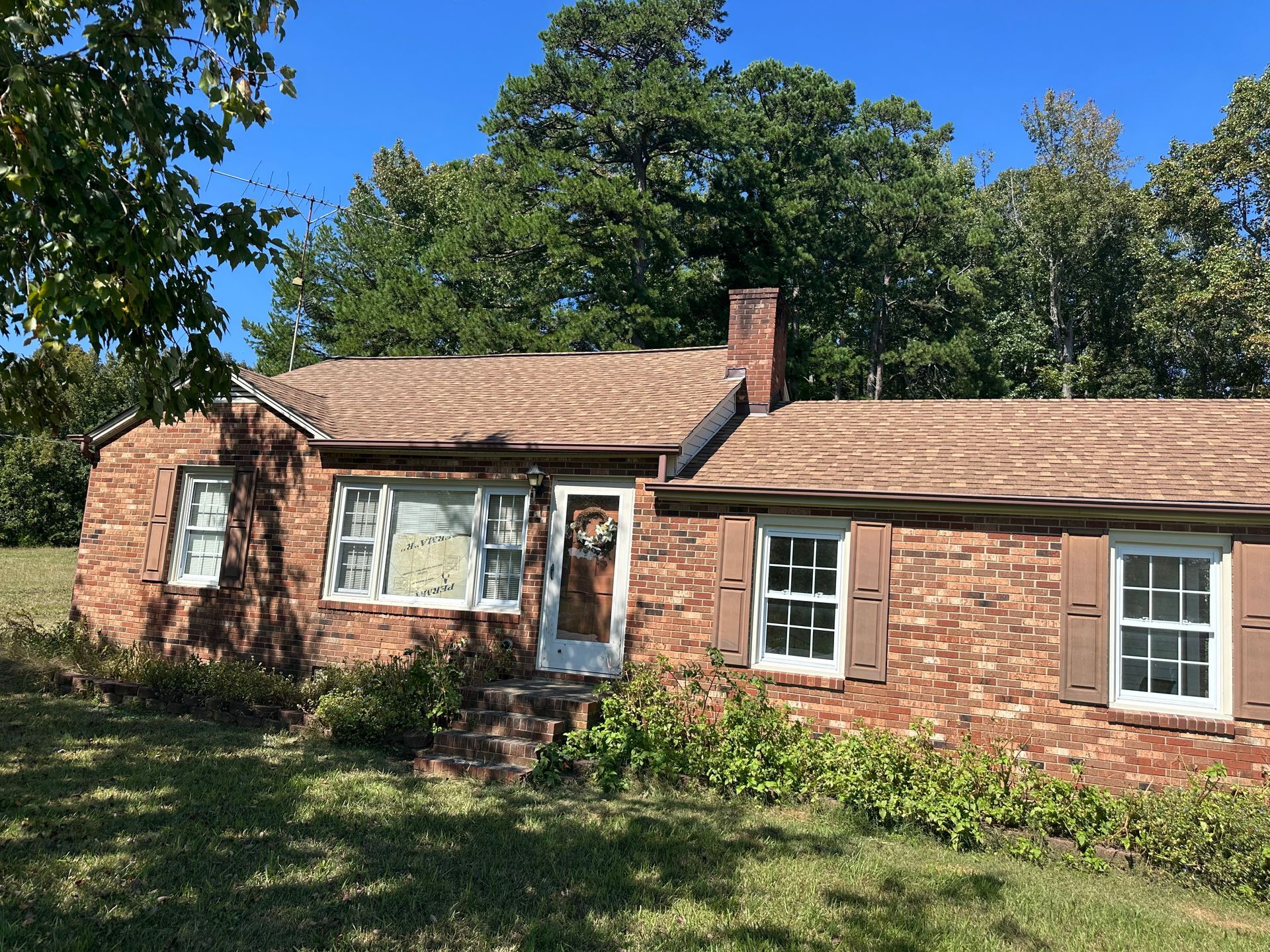 A brick house with a brown roof and white shutters is sitting in the middle of a grassy field.