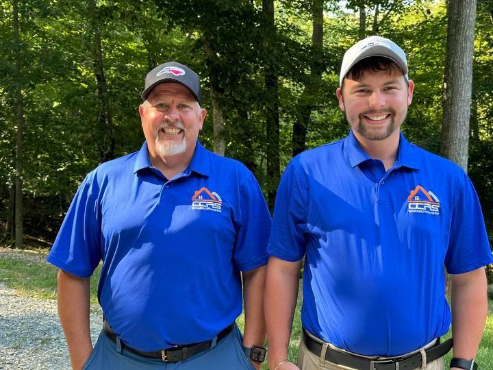 Two men in blue shirts are standing next to each other in front of trees.