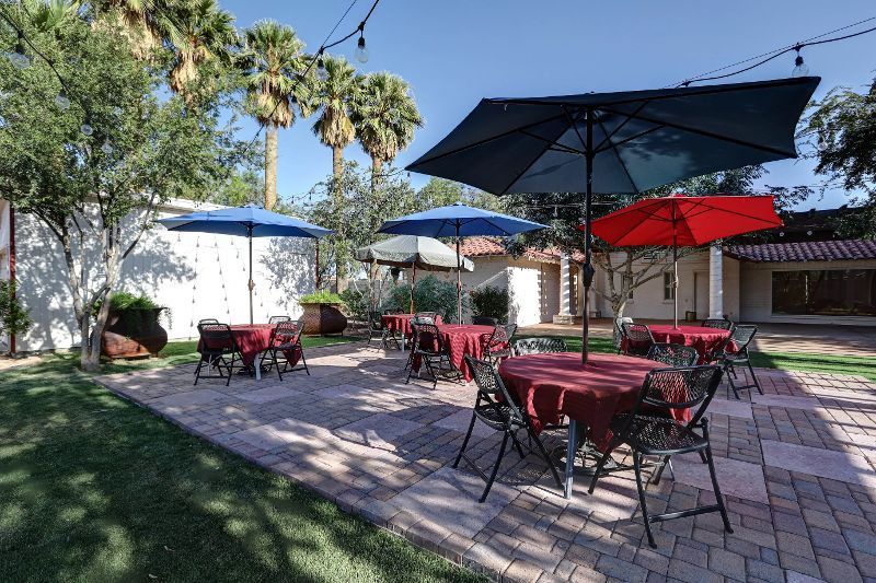Outdoor dining area with tables, red tablecloths, umbrellas, brick patio, and palm trees.