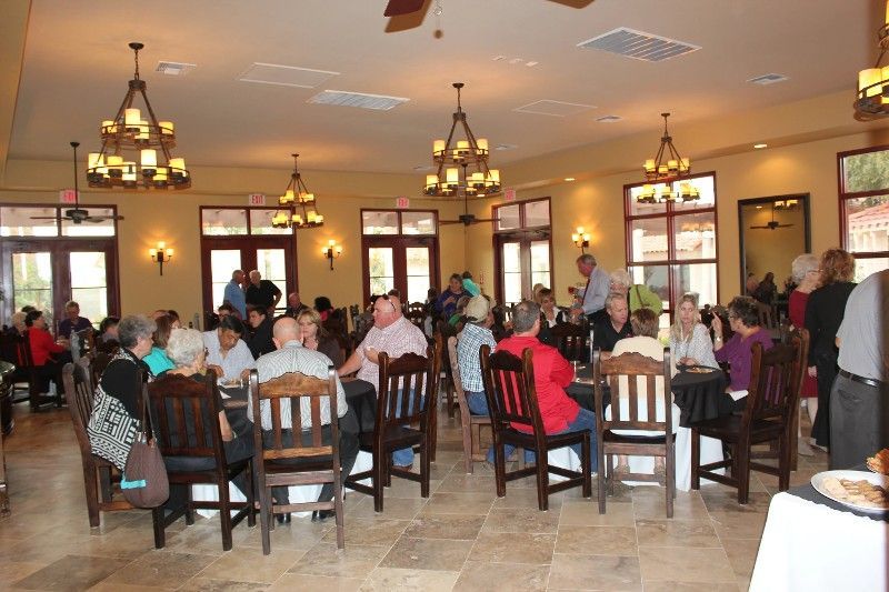 People seated at tables in a dining room, attending a gathering. Ornate chandeliers hang above.