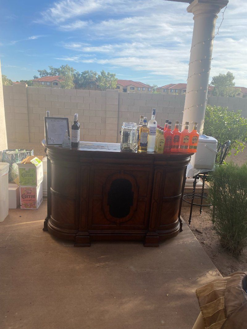 A wooden outdoor bar with liquor bottles on top. A backyard setting, sunny day.