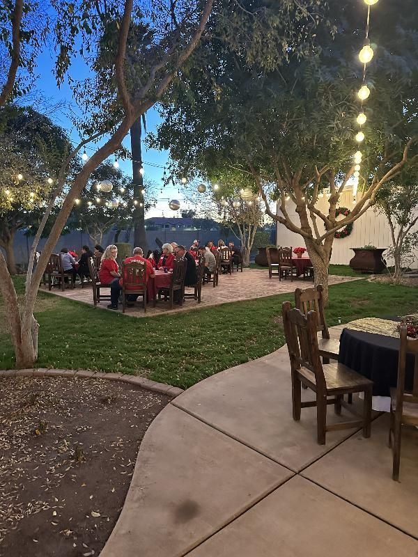 Outdoor dinner party at dusk, tables set with red tablecloths, string lights, people seated.