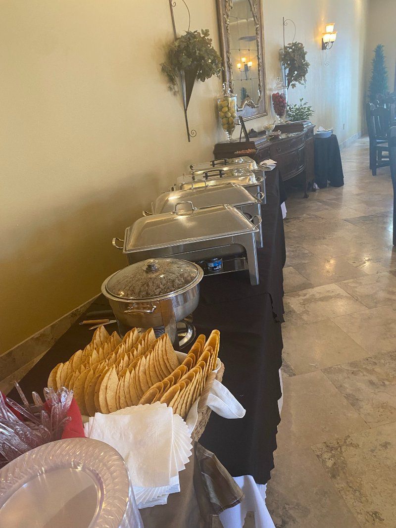Buffet table with silver chafing dishes, serving spoons, and stacked plates in a light-colored room.