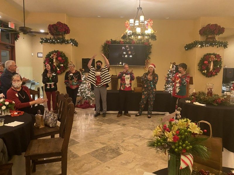 Group in holiday sweaters at a party, some singing. Decorated room with wreaths and Christmas tree.