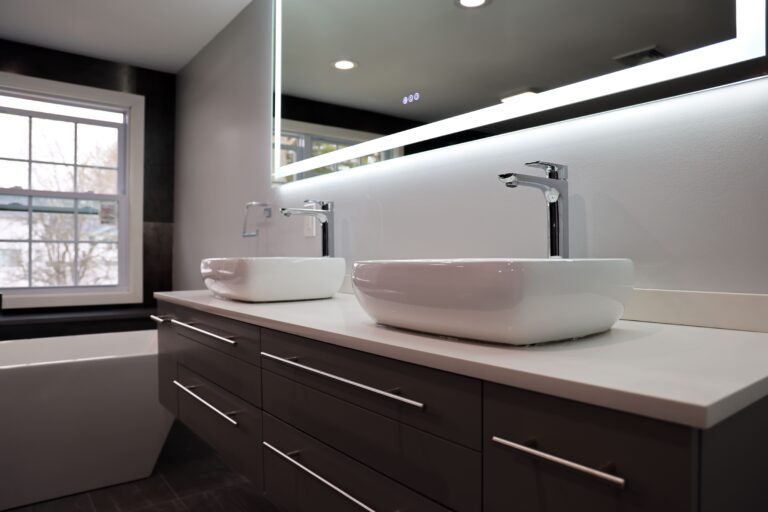 Modern bathroom vanity with two white vessel sinks, dark cabinets, mirror, and window light