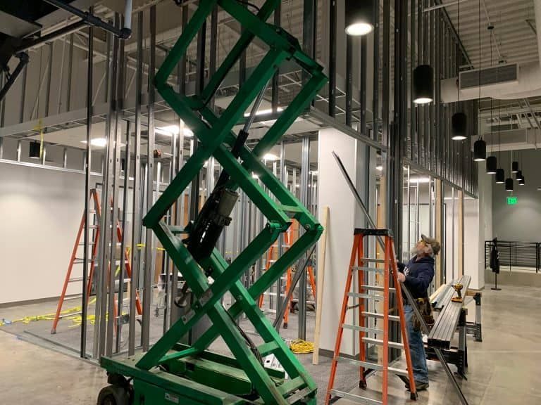 Green scissor lift being installed indoors, with ladders and a person working nearby under exposed ceiling lights