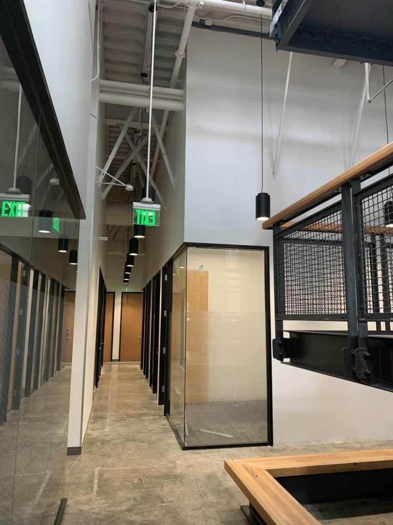 Industrial hallway with exposed ceiling, green EXIT signs, and a glass-walled room on the right.