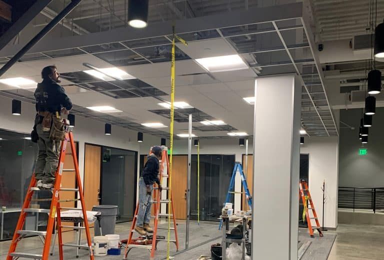 Workers on ladders installing ceiling fixtures in a large, unfinished commercial interior.