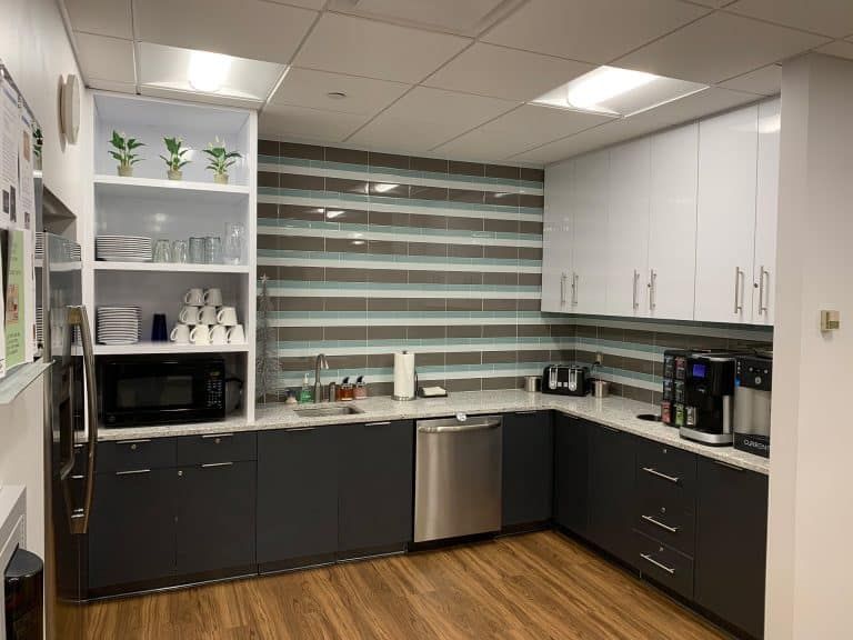 Modern office break room with white cabinets, dark counters, and striped window blinds