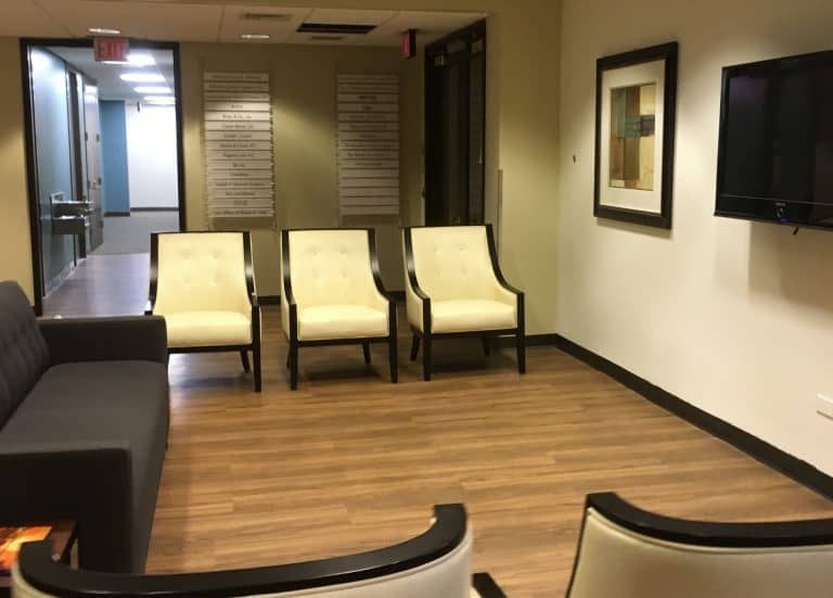 Empty waiting room with beige chairs, dark sofa, wood floor, wall-mounted TV, and hallway doors.