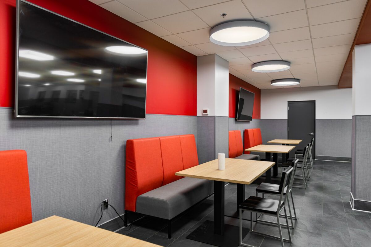 Modern café seating area with red booths, wooden tables, black chairs, and wall-mounted TVs