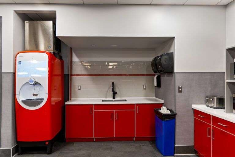 Red vending machine beside a red sink cabinet in a modern break room with a blue trash bin.