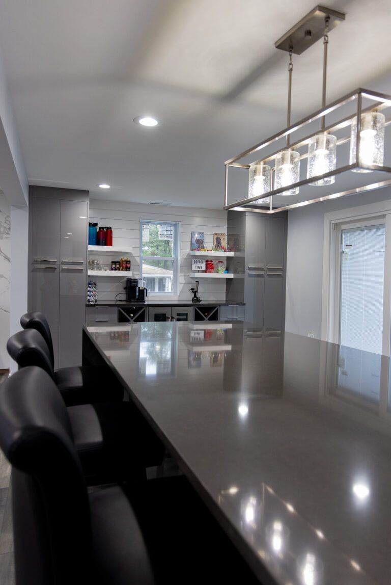 Modern kitchen with a reflective island, black stools, white cabinets, and a rectangular pendant light.