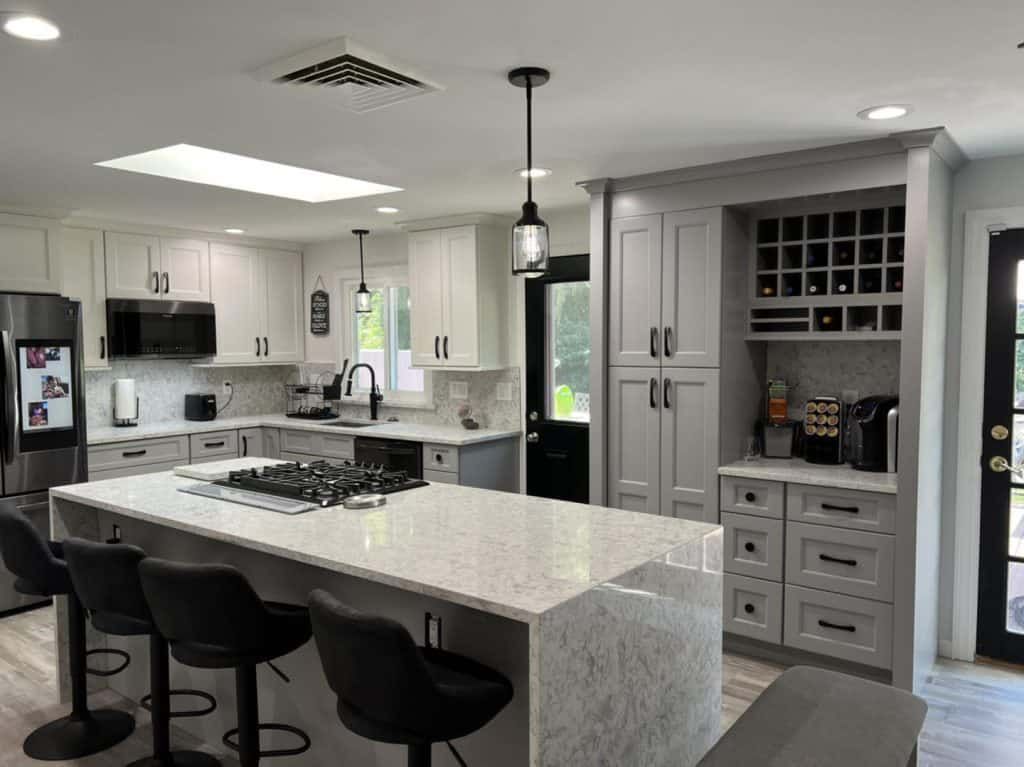 Modern white kitchen with large island, pendant lights, gray cabinets, and black bar stools.