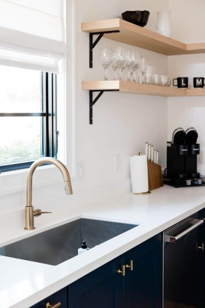 Modern kitchen with white countertops, black cabinets, brass faucet, and floating wood shelves with glassware