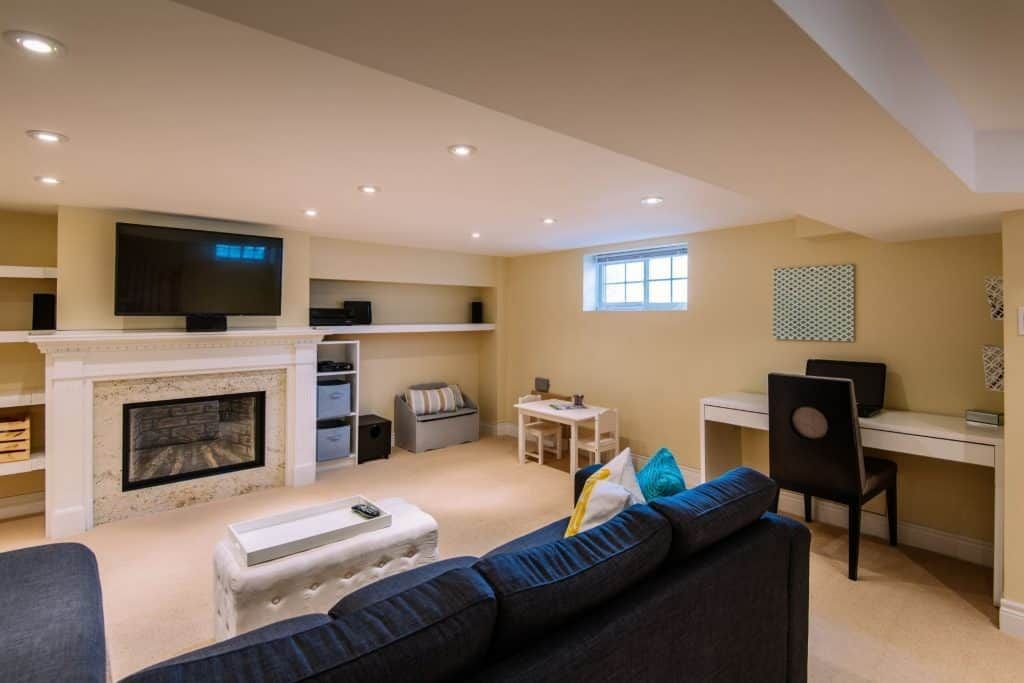 Cozy basement living room with fireplace, TV, desk, and dark sofa in beige decor
