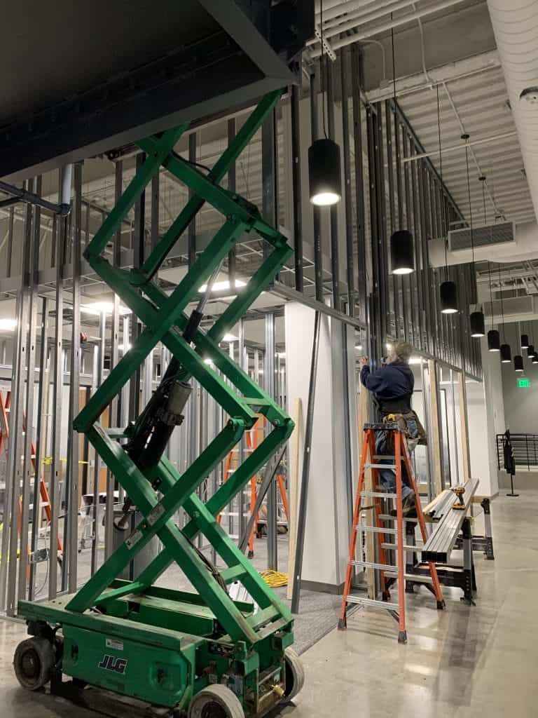 Green scissor lift in an industrial hallway beside ladders and hanging cables