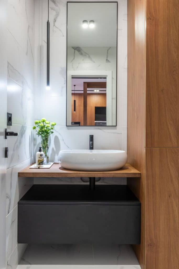 Narrow modern bathroom with wall mirror, white vessel sink, wood vanity, and black cabinet below