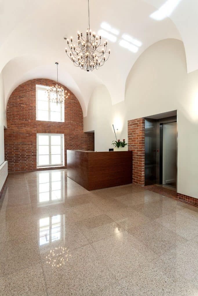 Spacious lobby with white vaulted ceiling, brick accent wall, chandelier, and polished terrazzo floor