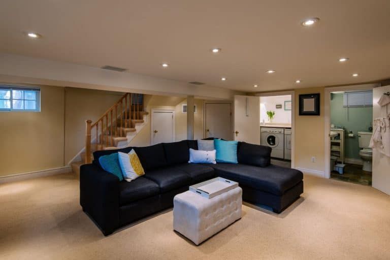 Spacious basement living room with black sectional sofa, ottoman, staircase, and recessed lighting.