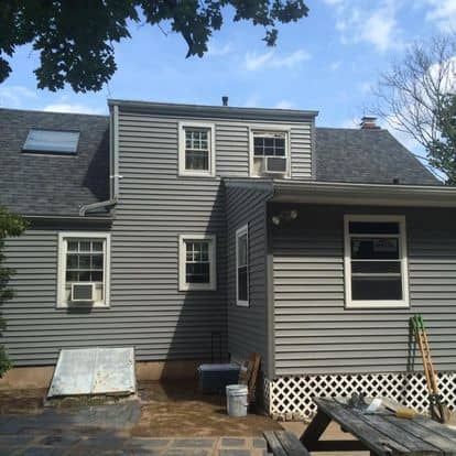 Gray house exterior with dormer windows, small side addition, and a low lattice porch under a blue sky