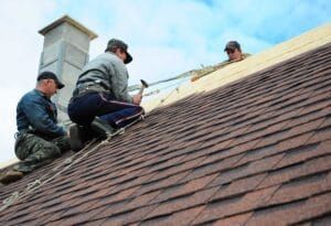 Three people work on a roof, installing shingles near a chimney.
