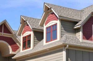 Gabled house with tan siding, red window frames, and red trim against a blue sky.