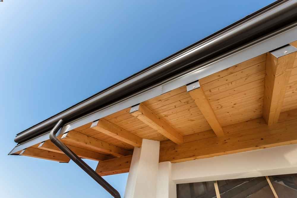 Wooden roof eaves with a dark gutter and downspout, against a blue sky.