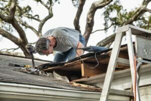 Man on roof repairing damage, using a tool. Ladder beside him.