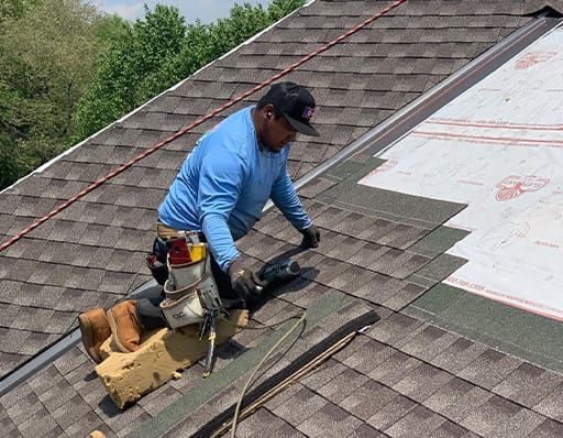 Roofer installing shingles on a roof. Blue shirt, black cap, tools in belt, sunlight.