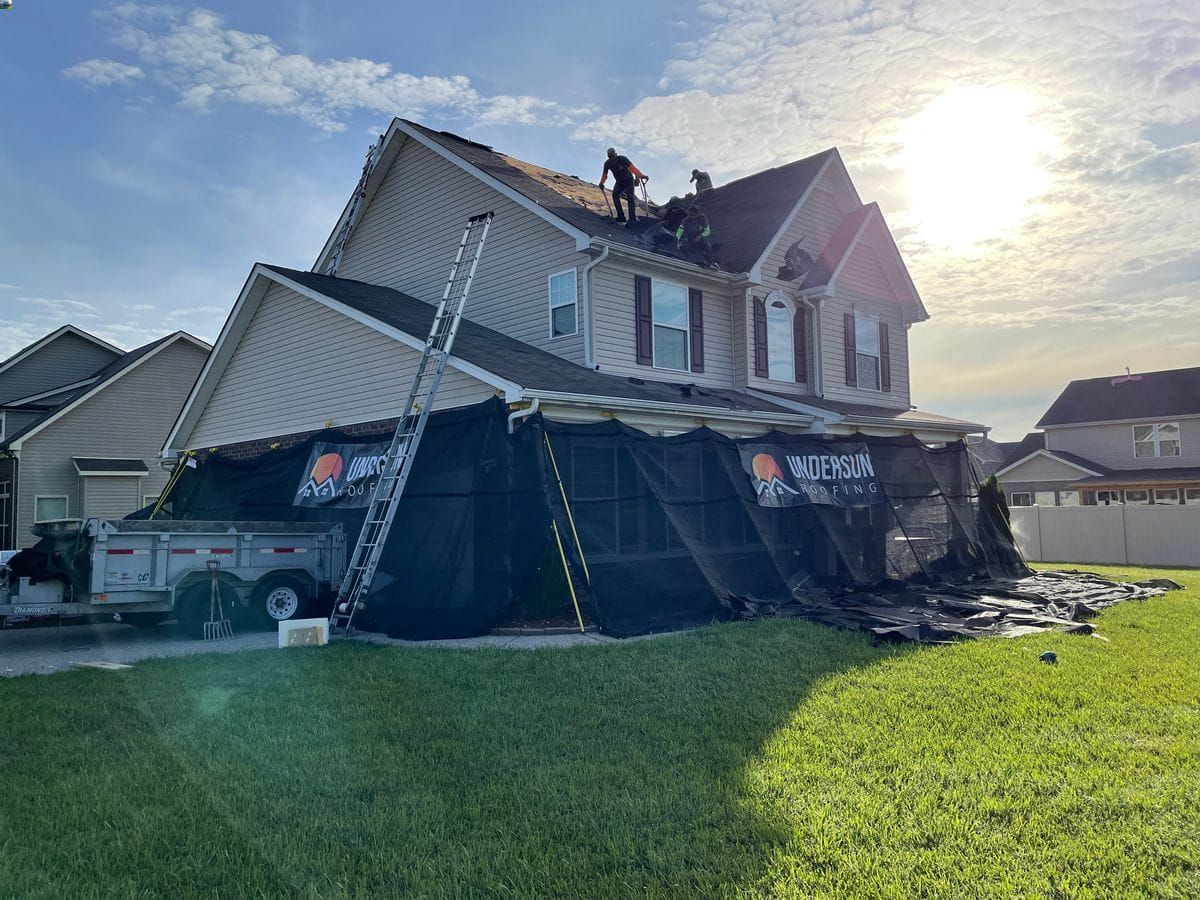 Two roofers on a house roof under repair, covered by black netting. A ladder rests against the house. Bright sunlight.