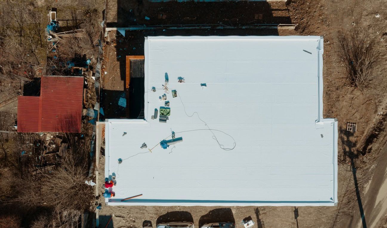 Overhead view of a building with a white roof, construction materials scattered across it, and a red-roofed structure nearby.