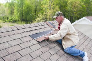 Man on roof replacing shingles. He wears a hat, and is kneeling on the roof with shingles in hand. Green trees are in the background.