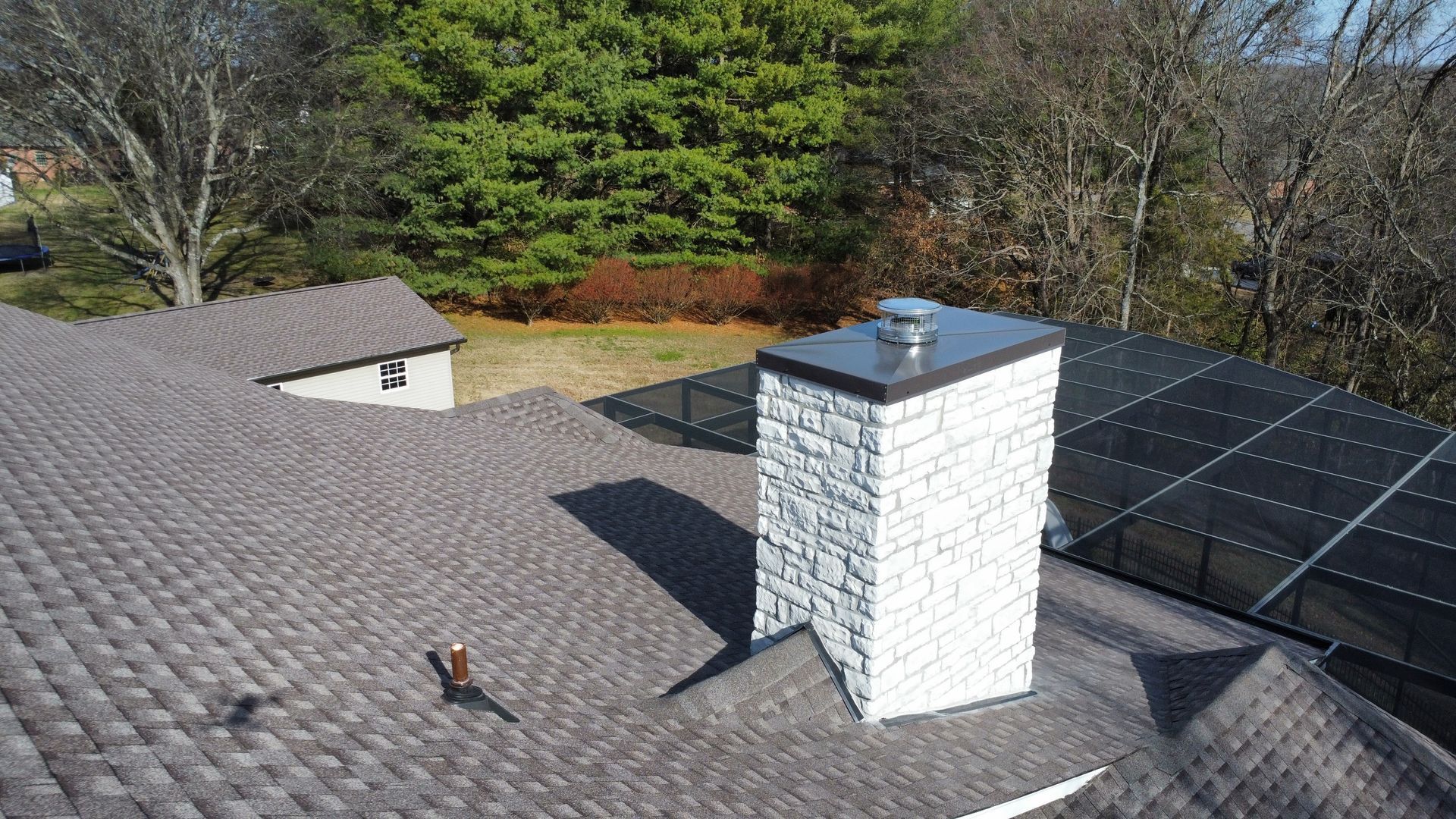 Rooftop with gray shingles, white brick chimney, and solar panels beside trees under a clear sky