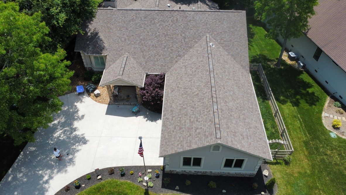 Aerial view of a suburban house with a gray roof, white driveway, and surrounding trees.