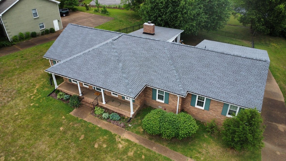 Aerial view of a single-story brick house with a gray roof, front porch, and surrounding lawn.
