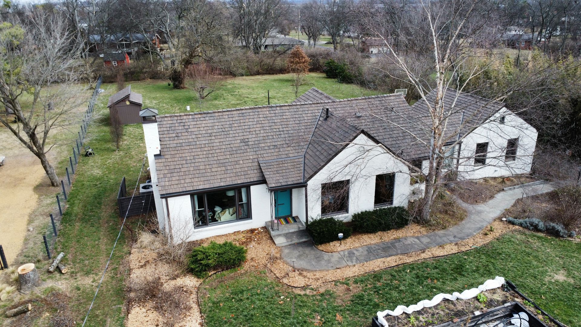 Aerial view of a small white house with a dark roof and a detached garage in a wooded yard.